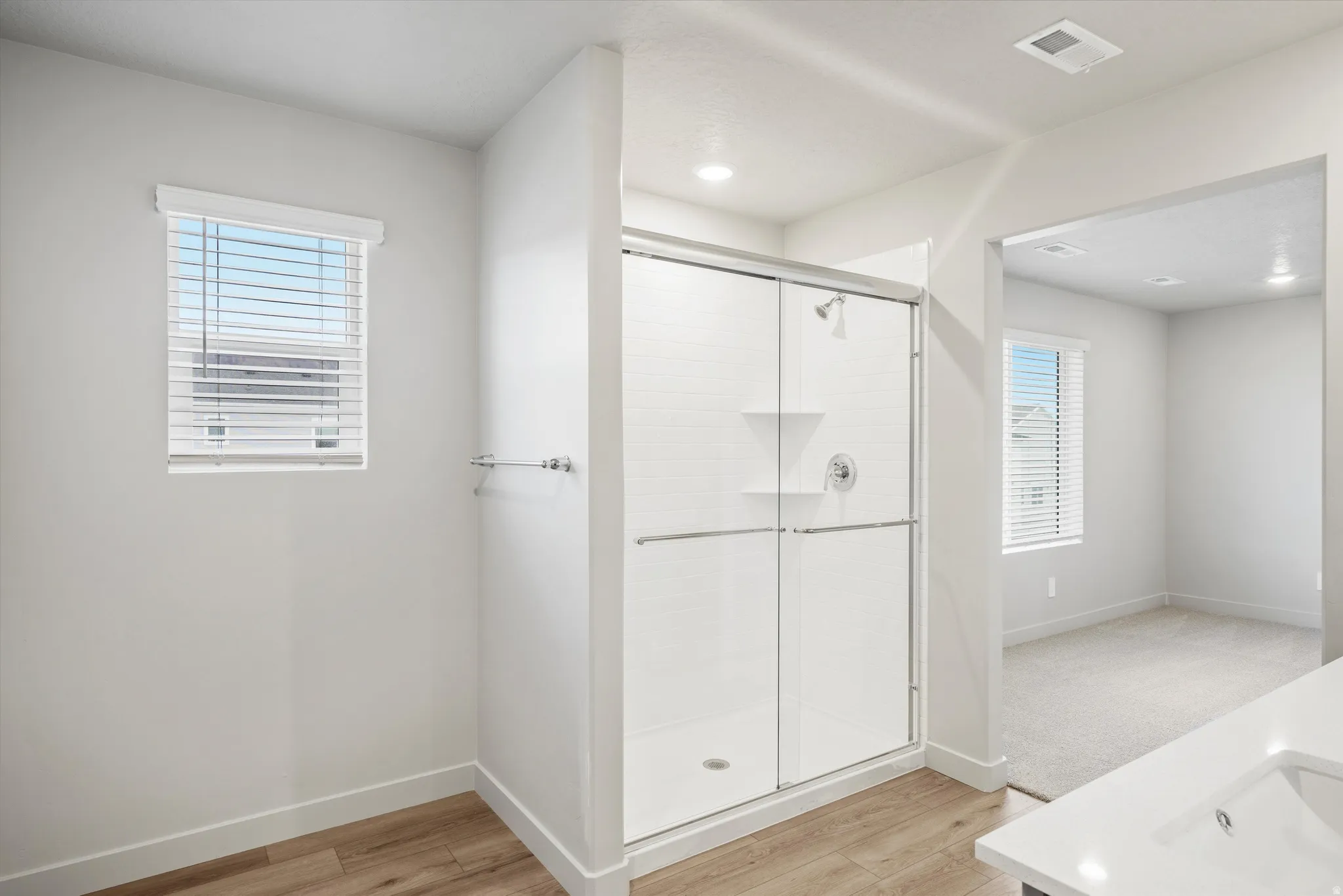 Spacious bathroom featuring a glass-enclosed shower with white subway tile surround, wood-finish flooring, and a window with white blinds
