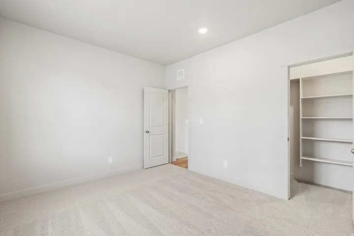 Spacious room featuring light-toned carpet flooring, light gray walls, a recessed ceiling light, and a built-in closet with shelving