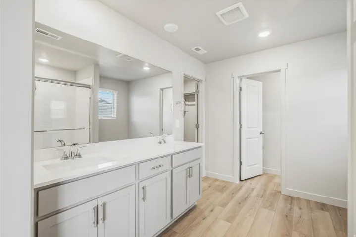 Bathroom featuring a double vanity with light gray cabinetry and white countertops, dual undermount sinks with chrome fixtures, a large frameless mirror, and light wood-finish flooring