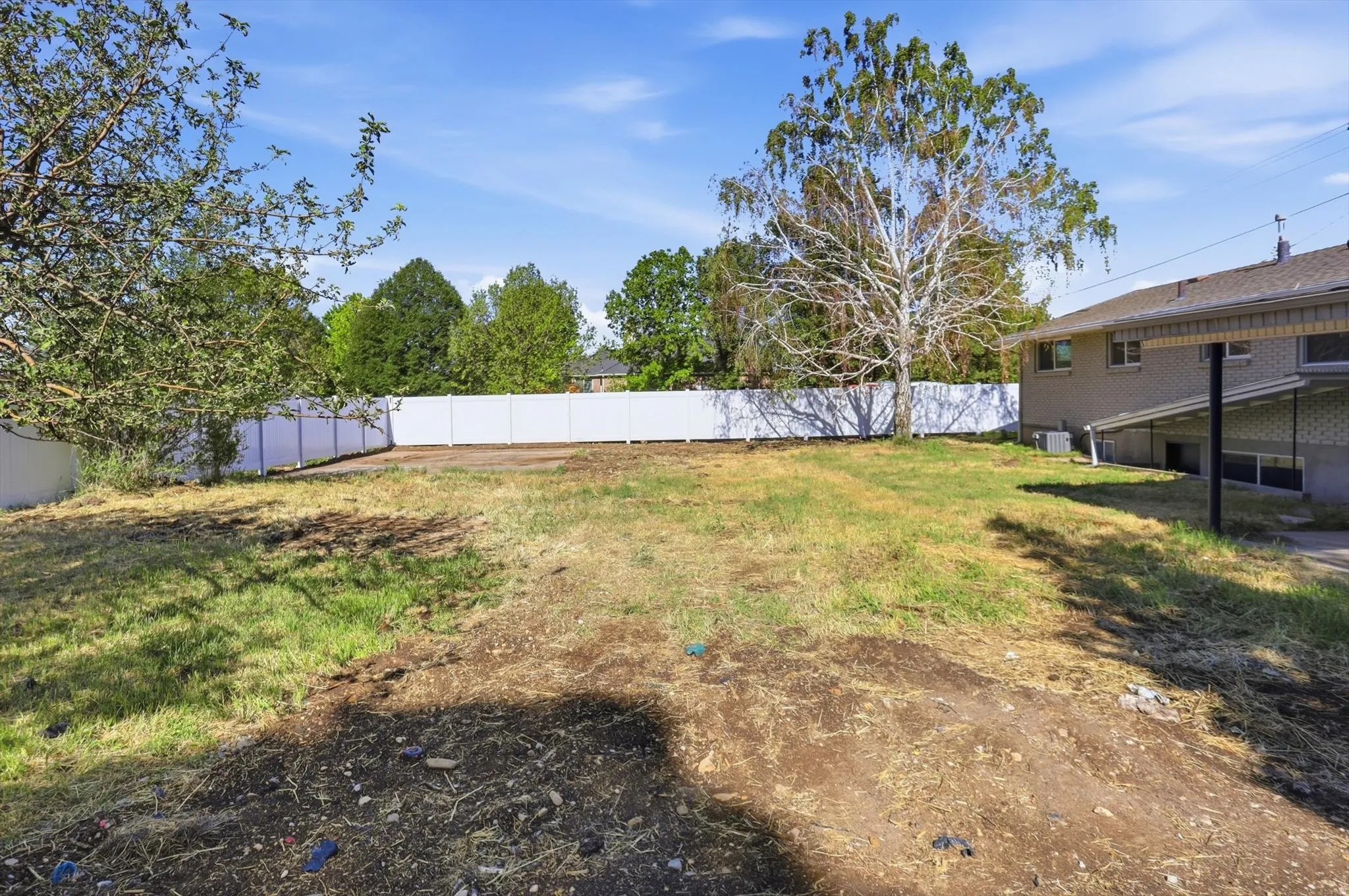 Expansive backyard featuring a white privacy fence, mature trees, and a partial brick exterior with a lower-level window