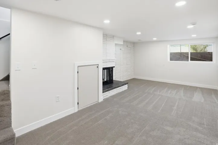 Spacious living area featuring light gray carpeting, recessed ceiling lighting, a brick fireplace with a dark stone hearth, a multi-pane window, and white painted walls