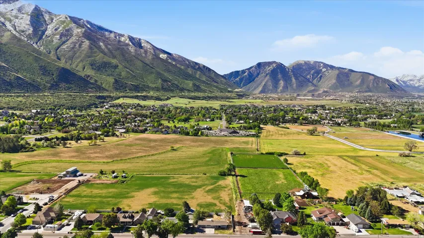 Expansive aerial perspective showcasing a mountainous backdrop, verdant fields, a residential area, and a visible body of water