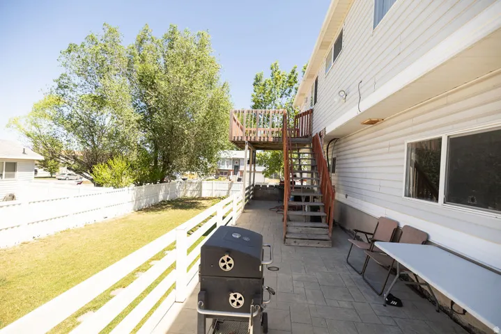 Ground-level patio with stamped concrete flooring, white picket fence, mature trees, and an exterior staircase leading to an upper-level wood deck