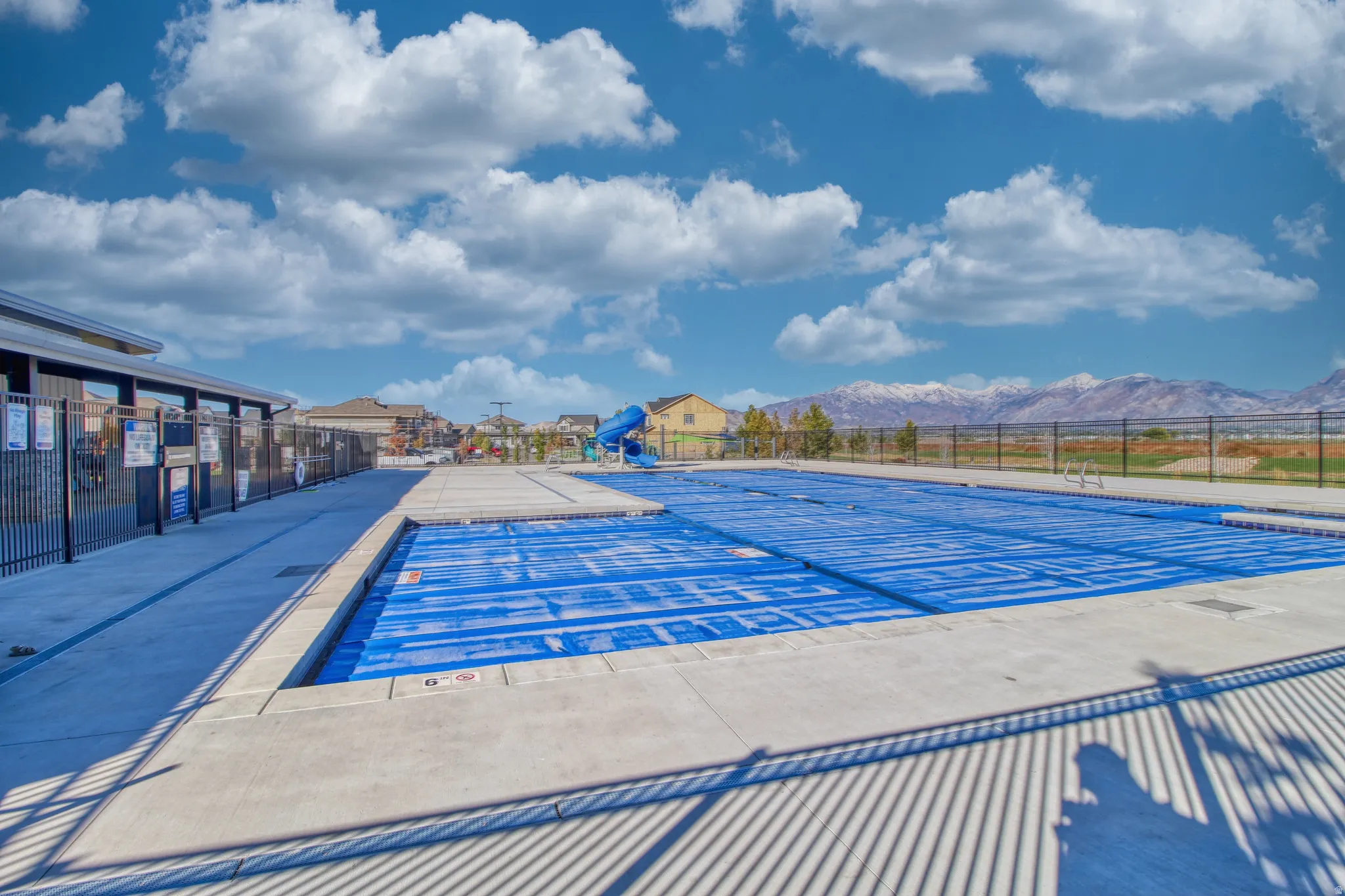 Community swimming pool with a blue cover, surrounded by concrete decking and a black metal fence