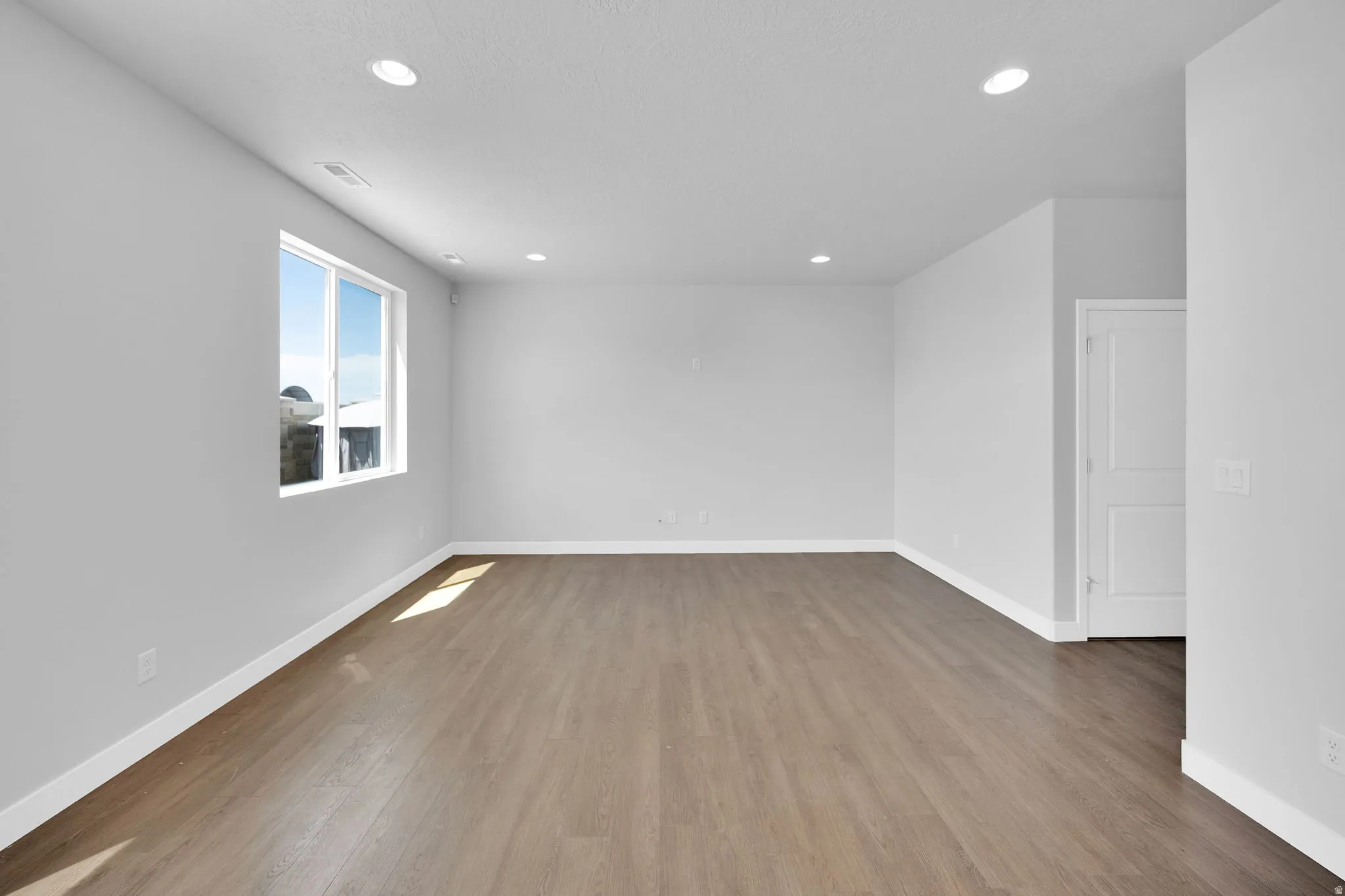 Spacious room featuring wood-finish flooring, recessed lighting, a large window, white baseboards, and a paneled door