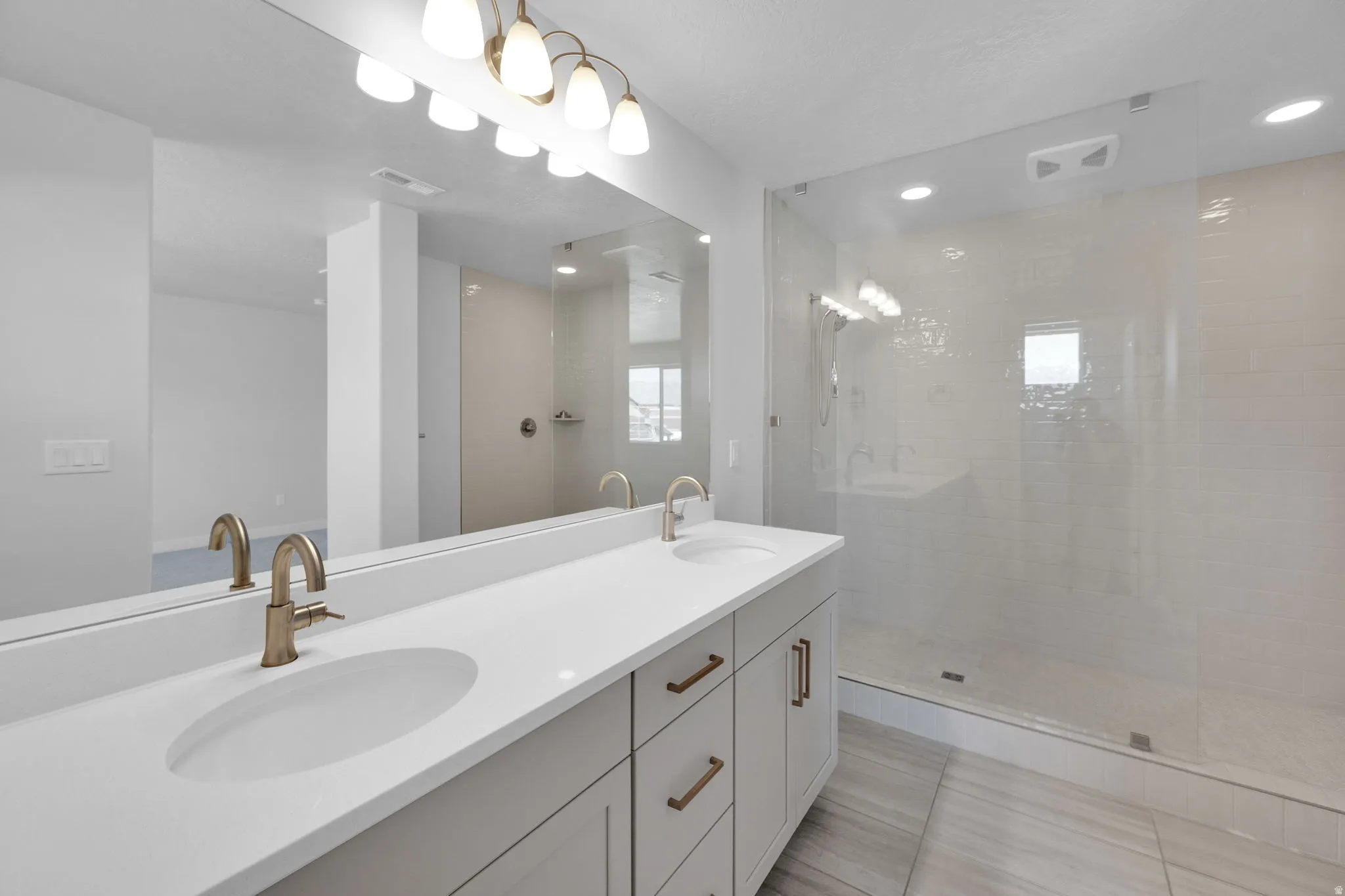 Dual vanity with undermount sinks and brushed gold fixtures, a full-width mirror, a frameless glass shower enclosure, a tiled shower interior, and recessed lighting