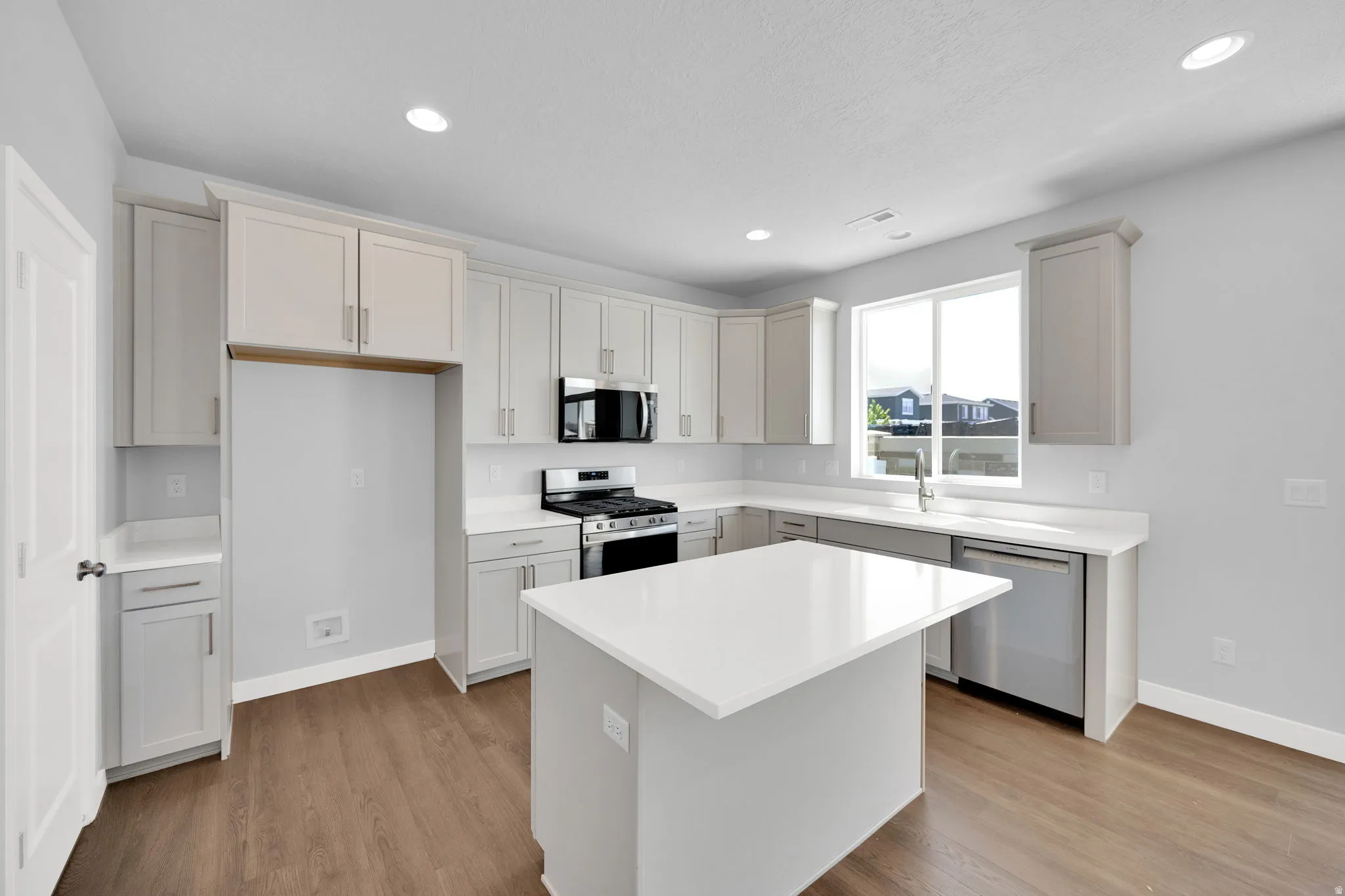 Modern kitchen featuring an island with a white countertop, light gray cabinetry, stainless steel appliances, wood-finish flooring, and recessed lighting