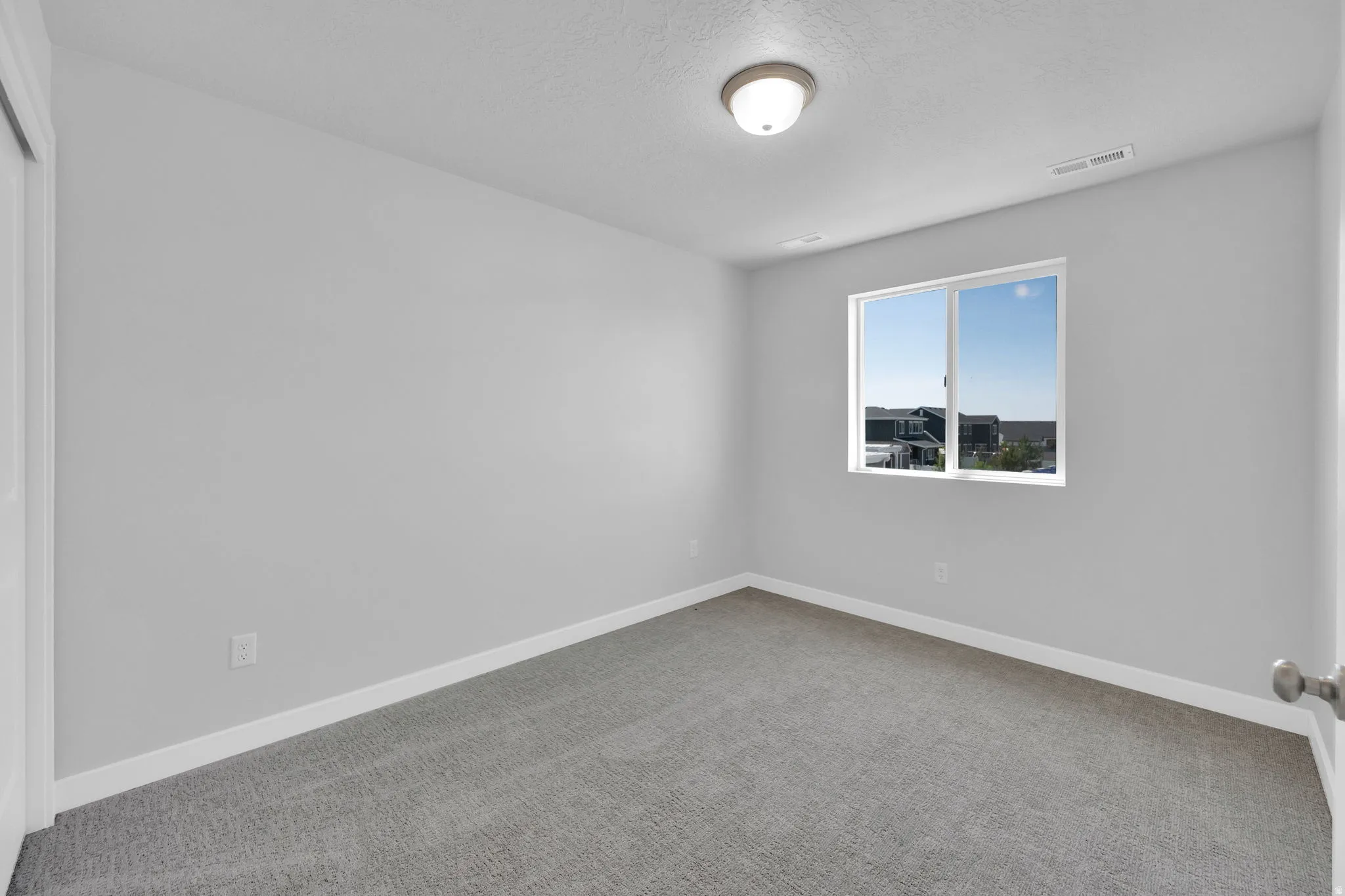 Bright interior room featuring a window with exterior residential structures visible, light gray wall paint, white trim, gray carpet flooring, and a flush-mount ceiling light fixture