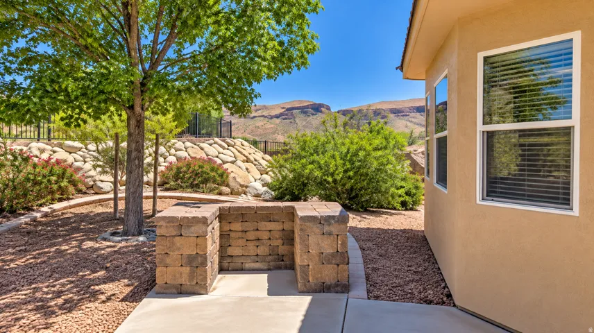 Stucco exterior with multiple windows, paver block outdoor living structure, mature landscaping, and a rock retaining wall