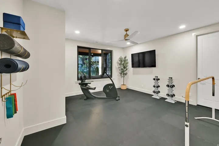 Dedicated exercise room featuring dark rubber flooring, a large window, a ceiling fan with integrated lighting, recessed lighting, and a wall-mounted display