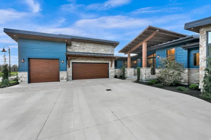 Modern exterior featuring blue horizontal siding, stone accents, two wood-finish garage doors, a covered entry with exposed wood beams, and a concrete driveway