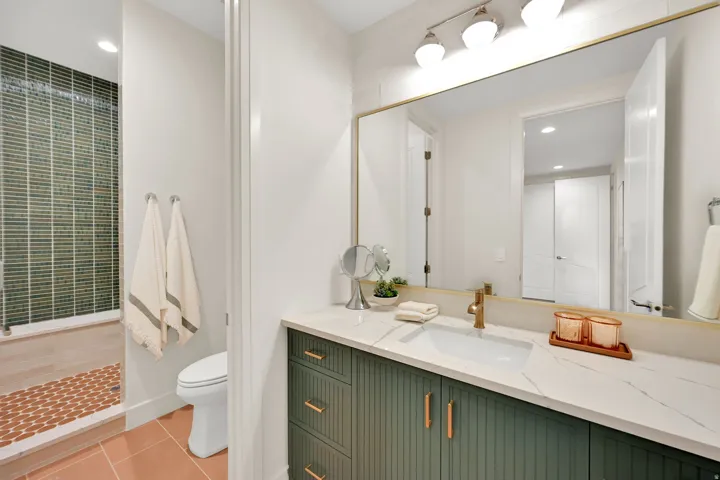 Bathroom featuring a green slatted vanity with copper hardware, a white countertop with an integrated rectangular sink, a gold-framed mirror, and a walk-in shower with green mosaic tile