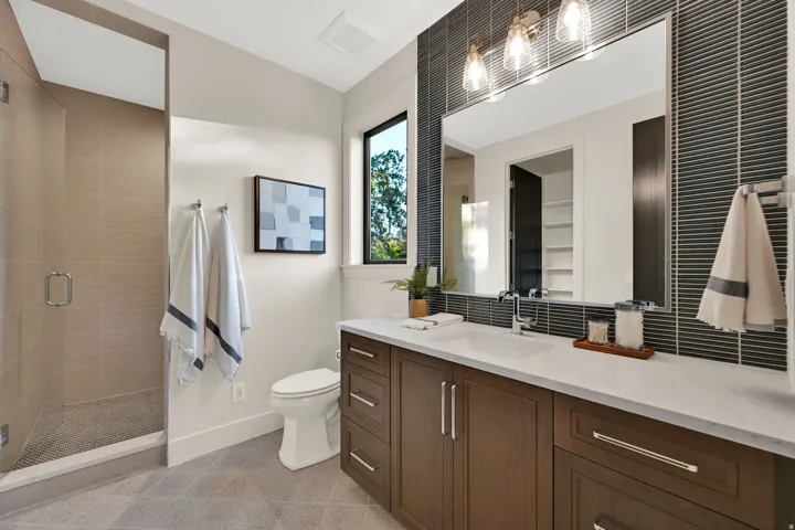 Contemporary bathroom featuring a spacious glass-enclosed shower with tile surround, a dark wood-finish vanity with ample storage, a white countertop, and a large mirror with integrated lighting