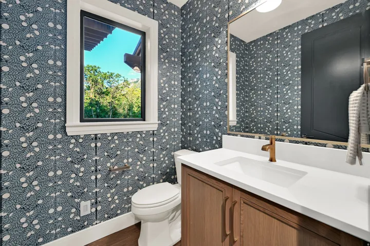 Powder room featuring botanical-patterned wallpaper, a single window with white trim, and a wood-finish vanity with a white countertop and integrated sink