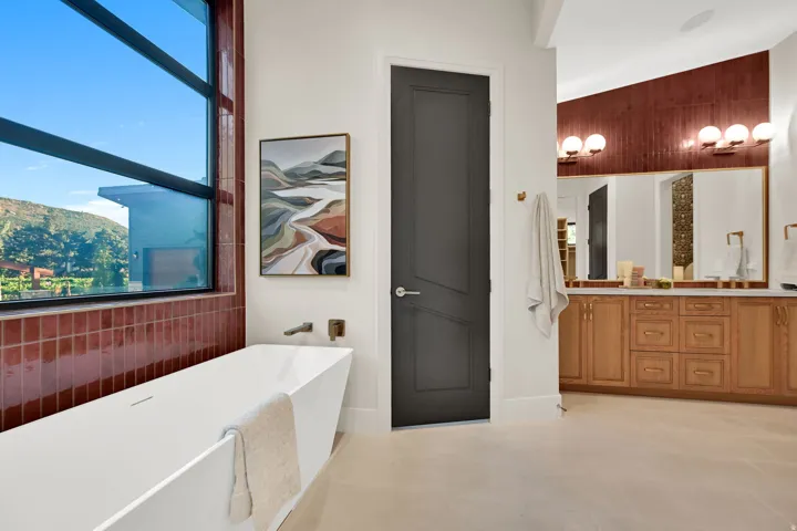 Contemporary bathroom featuring a freestanding tub, large window, wood-finish vanity with dual sinks, and a textured accent wall with wall-mounted sconces