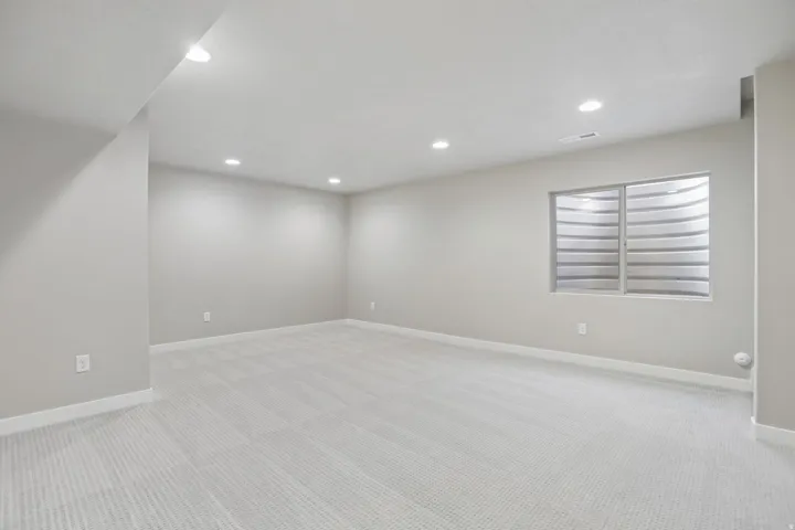 Spacious interior room featuring light-colored carpeting, recessed ceiling lighting, a window with a well-cover, and neutral wall tones