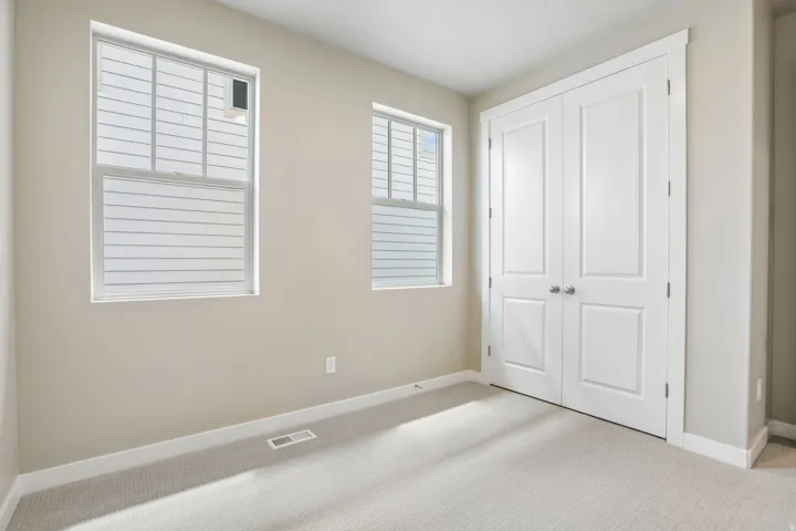 Neutral tone wall paint with white trim, two double-hung windows, light-colored carpeting, and a double-door closet with panel detailing