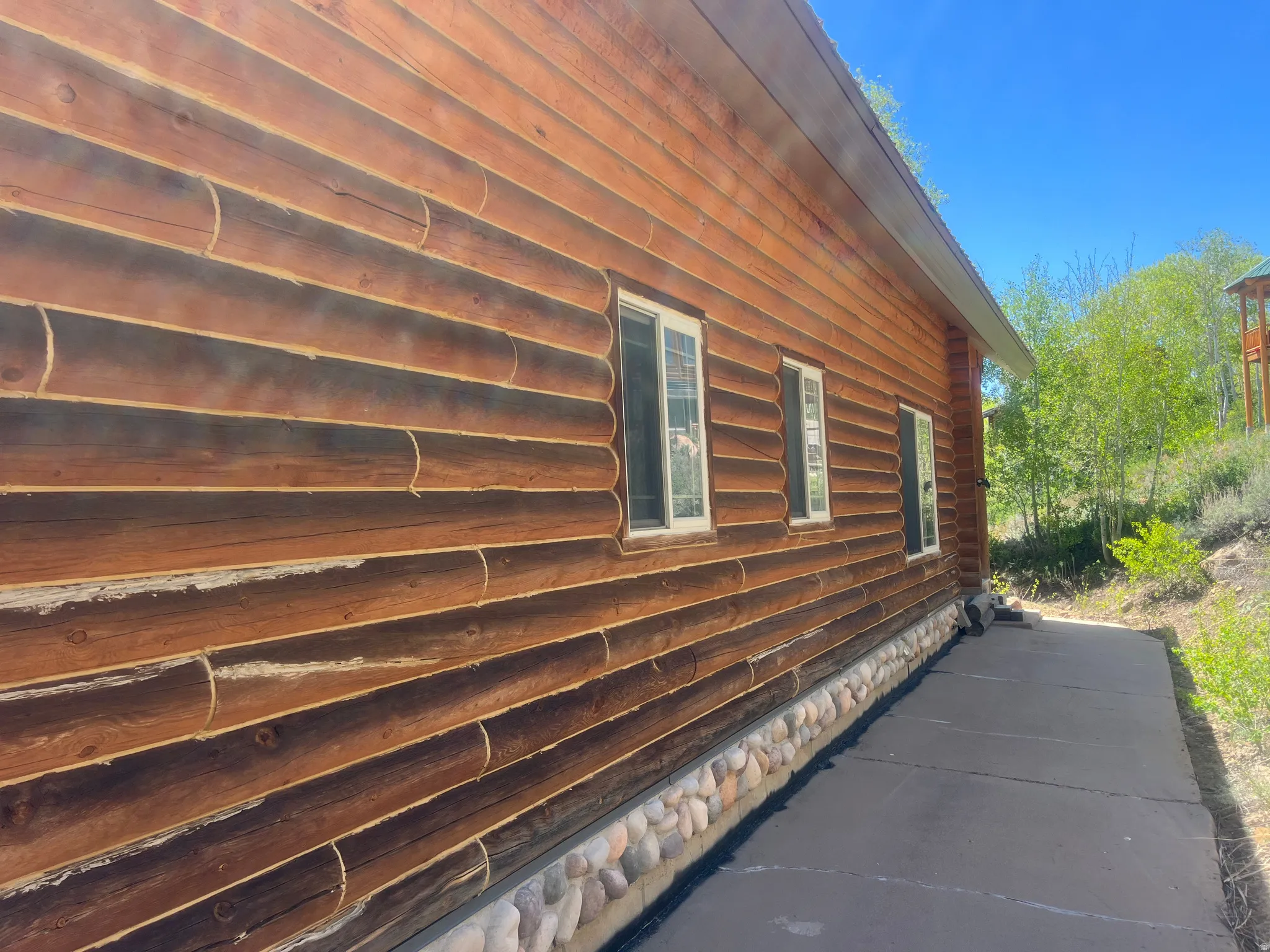 Log-style exterior siding featuring white-framed windows, a stacked stone foundation, and a dark paved walkway