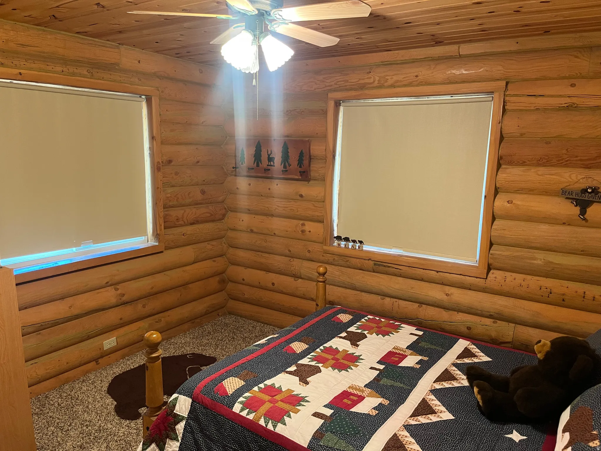 Interior log walls and ceiling, featuring two windows with roller shades, an overhead ceiling fan with light fixture, and carpet flooring