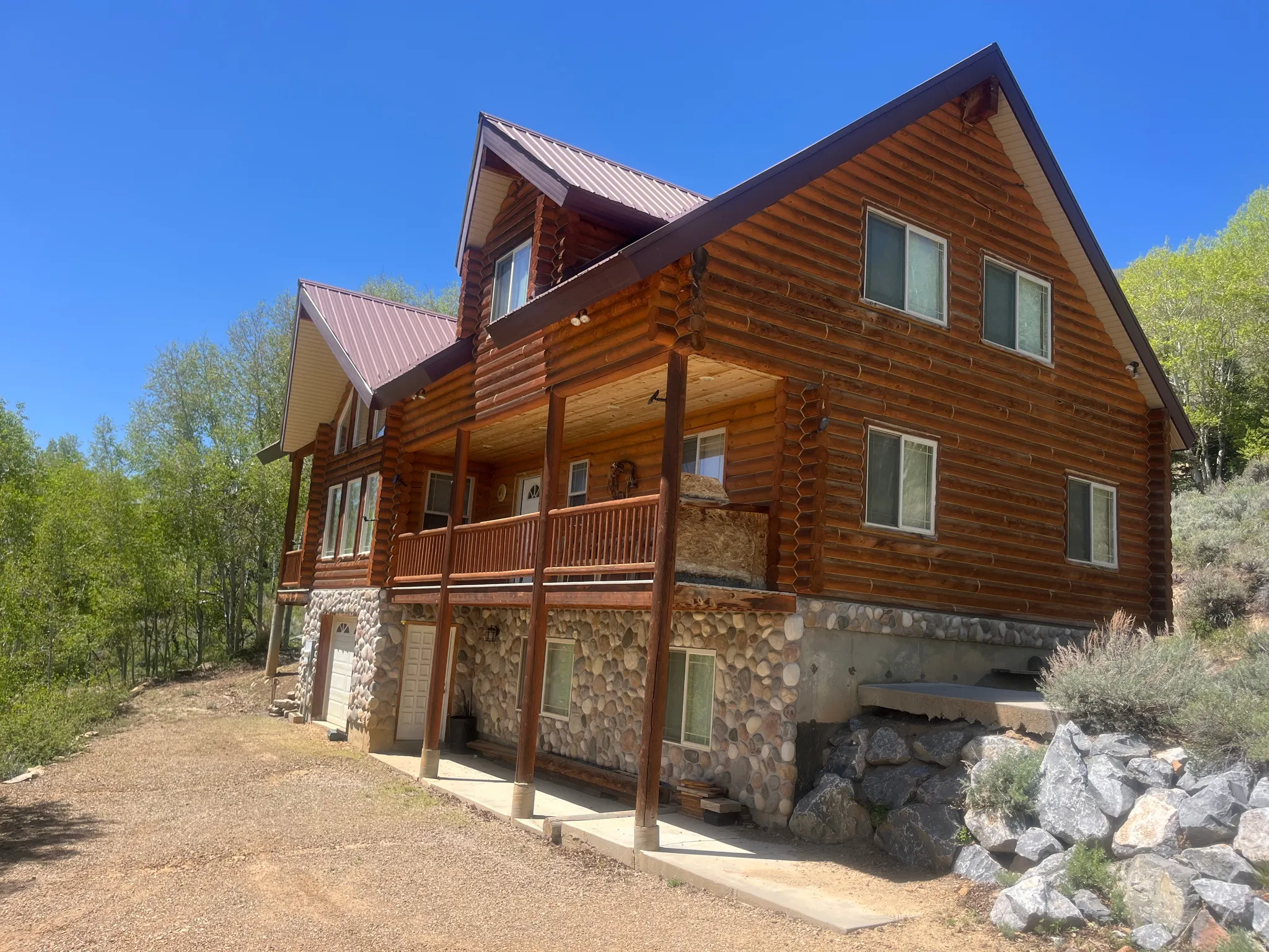 Log cabin exterior featuring a brown metal roof, stone foundation, multi-level covered porch, and exposed wood siding