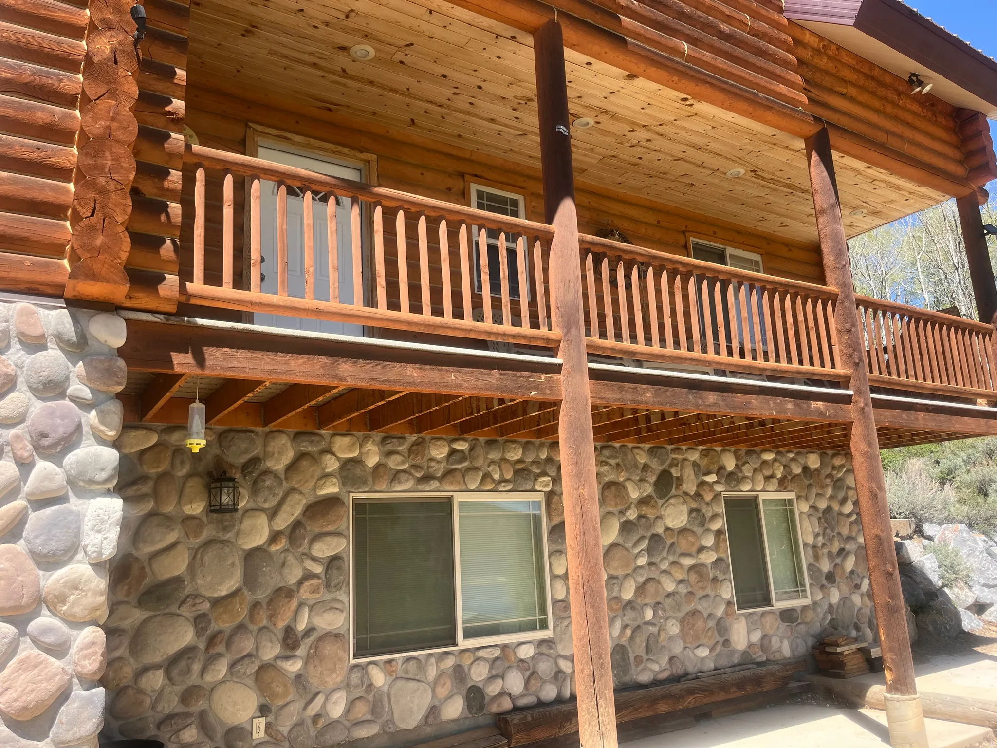 Exterior featuring log construction, a full-length upper-level balcony with wood railing, a lower-level stone facade, and multiple windows