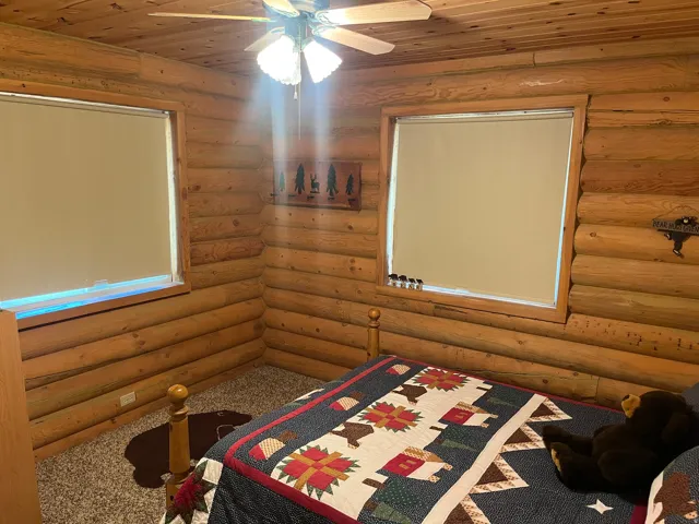 Interior log walls and ceiling, featuring two windows with roller shades, an overhead ceiling fan with light fixture, and carpet flooring