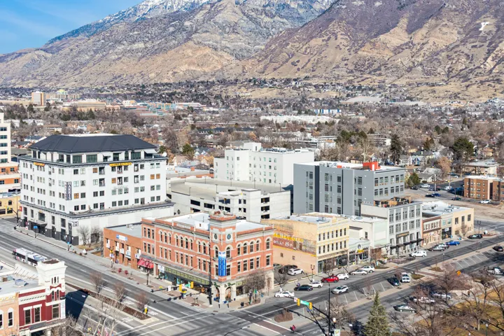 Urban landscape featuring diverse architectural styles, a prominent multi-story building with a dark roof, and a backdrop of mountains