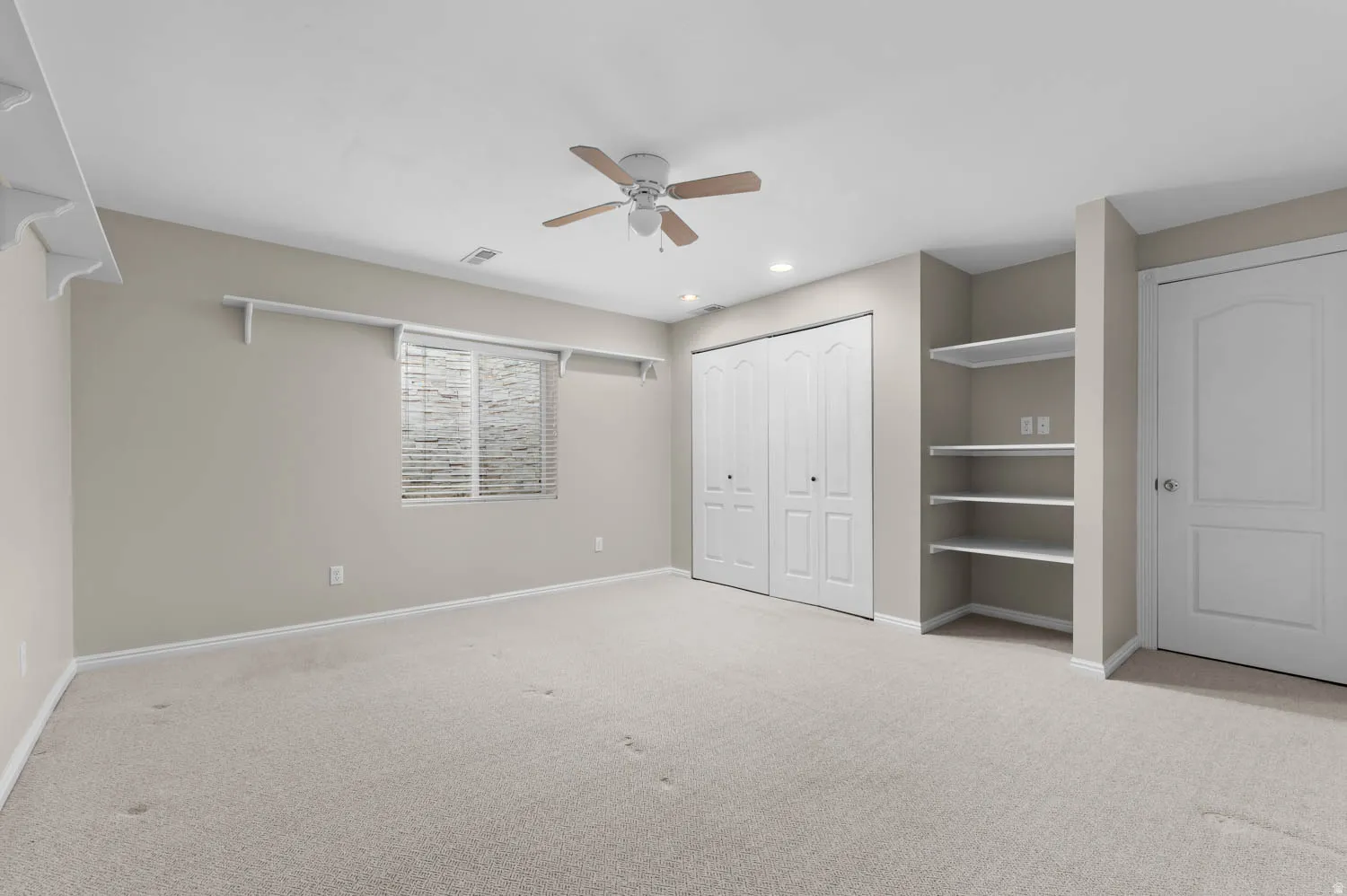 Spacious room featuring neutral carpet flooring, a ceiling fan, a window with horizontal blinds, built-in shelving, and bi-fold closet doors
