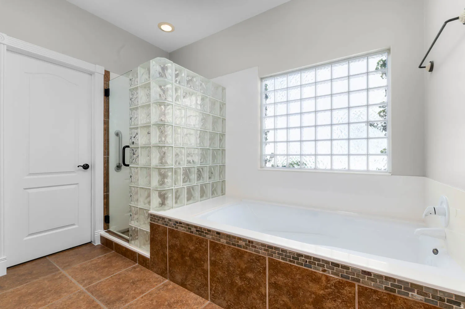 Bathroom featuring a glass block shower enclosure, a white soaking tub with tiled surround, a large glass block window, and brown tile flooring