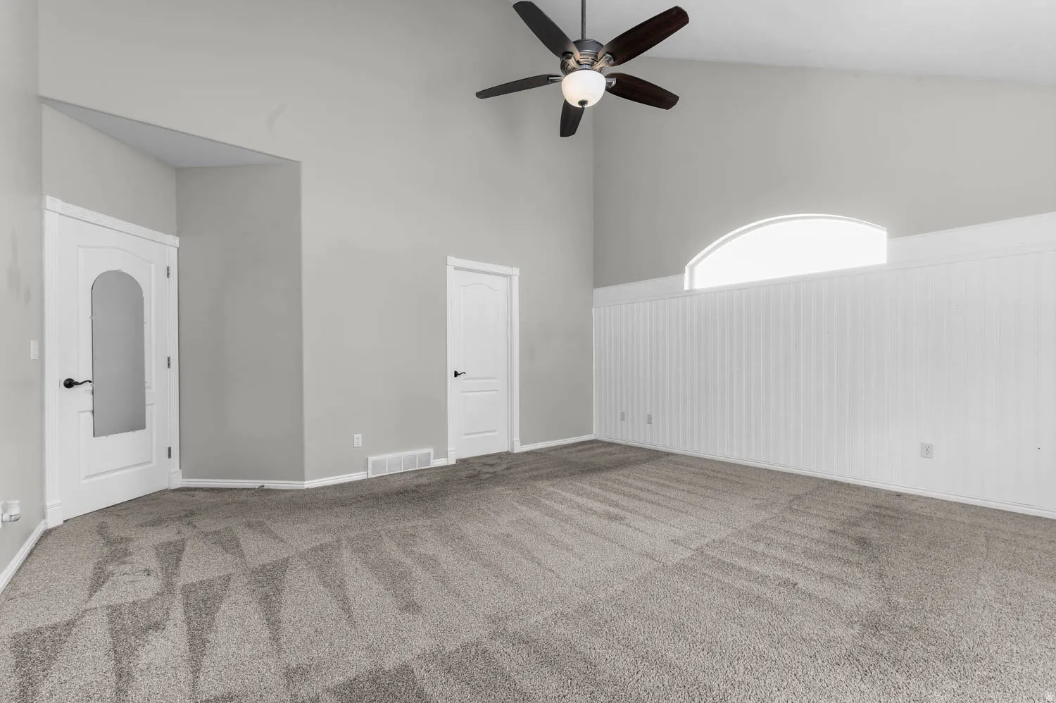 Vaulted ceiling room featuring light gray walls, a ceiling fan with integrated lighting, and a prominent arched window above white beadboard paneling