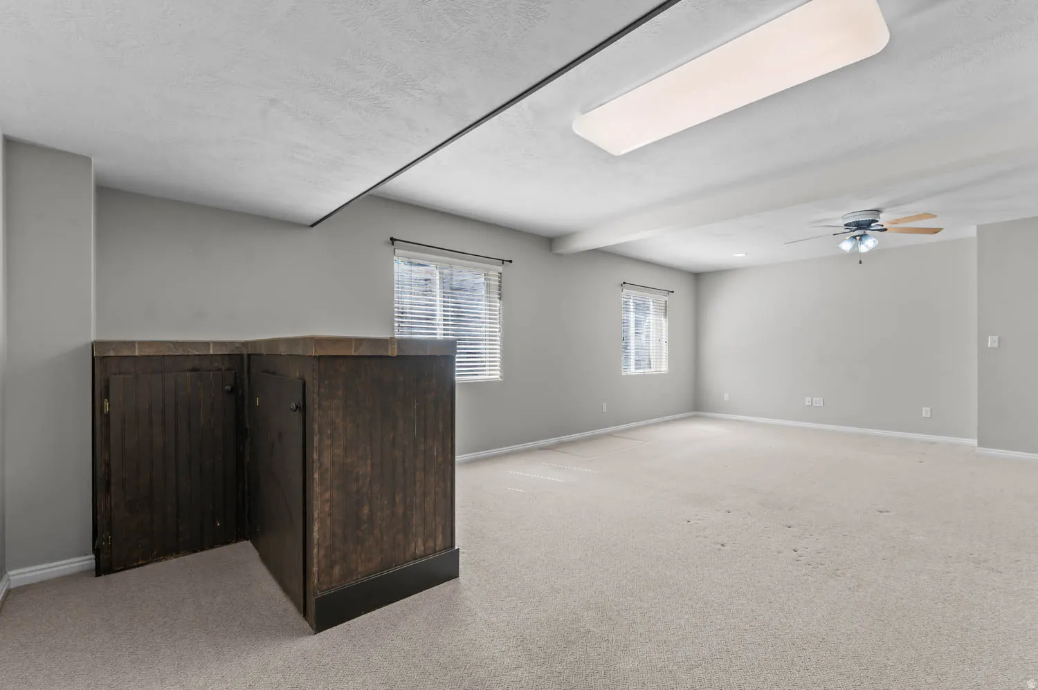 Spacious room featuring light-toned carpeting, two windows with blinds, and a built-in dark wood-finish cabinet with a tiled countertop