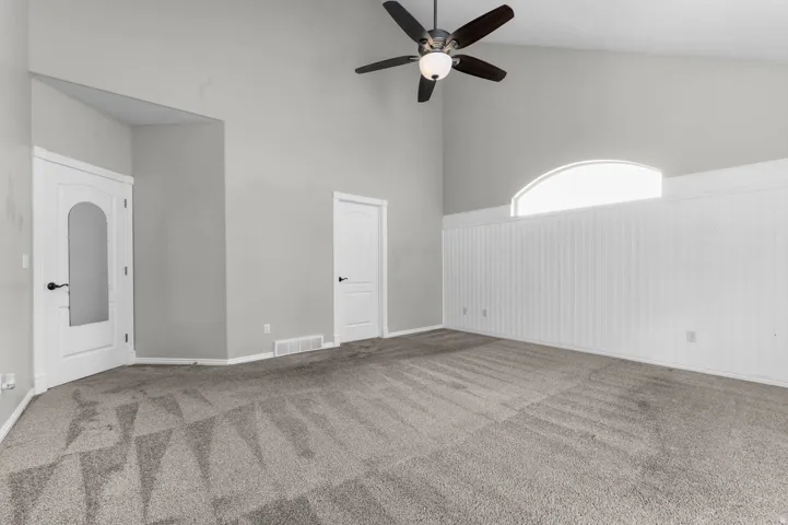 Vaulted ceiling room featuring light gray walls, a ceiling fan with integrated lighting, and a prominent arched window above white beadboard paneling