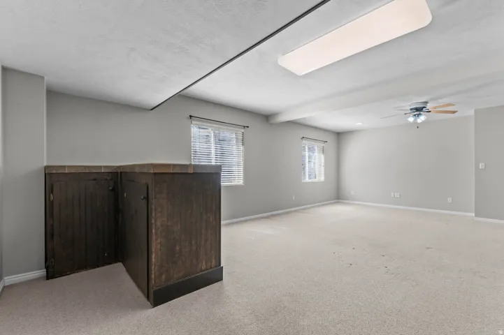Spacious room featuring light-toned carpeting, two windows with blinds, and a built-in dark wood-finish cabinet with a tiled countertop