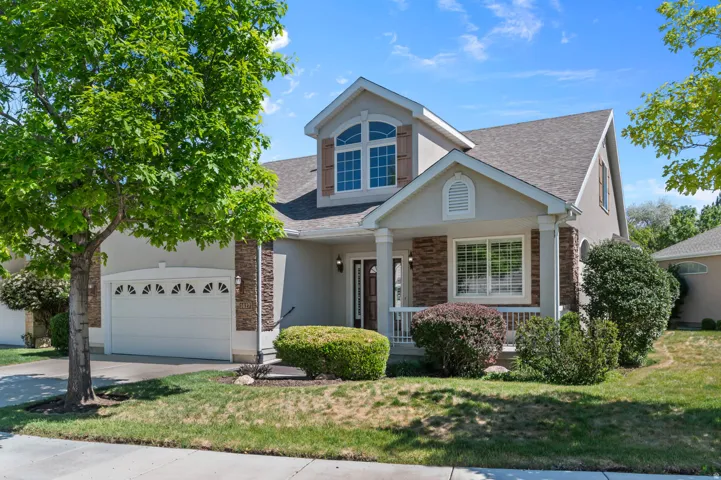 Stucco facade residence featuring a covered front porch with white railing, stone accents, arched upper window with shutters, and an attached two-car garage with decorative window inserts