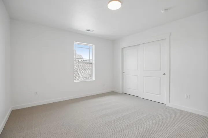 Neutral-toned carpeting extends throughout this room, complemented by a window with white trim, a built-in closet with sliding panel doors, and a flush-mount ceiling light fixture