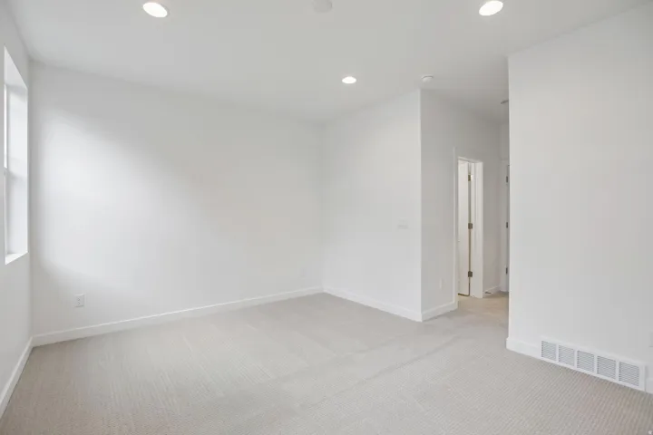 Spacious interior room featuring light gray carpeting, crisp white walls, recessed ceiling lighting, and a prominent window providing natural light