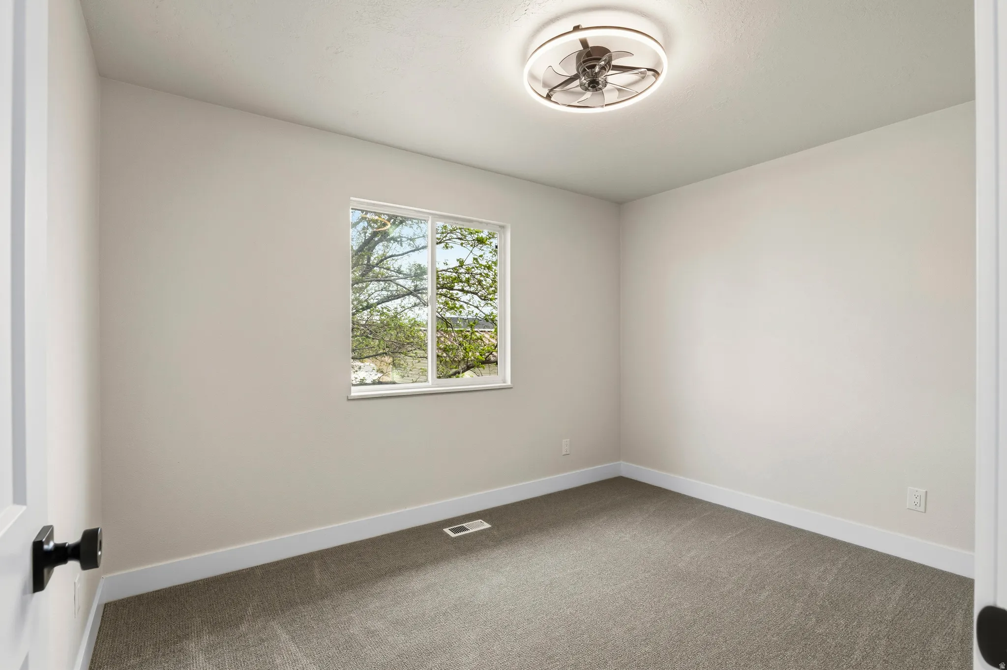Bright room featuring a window overlooking greenery, plush gray carpeting, crisp white baseboards, and a contemporary ceiling fan with integrated lighting
