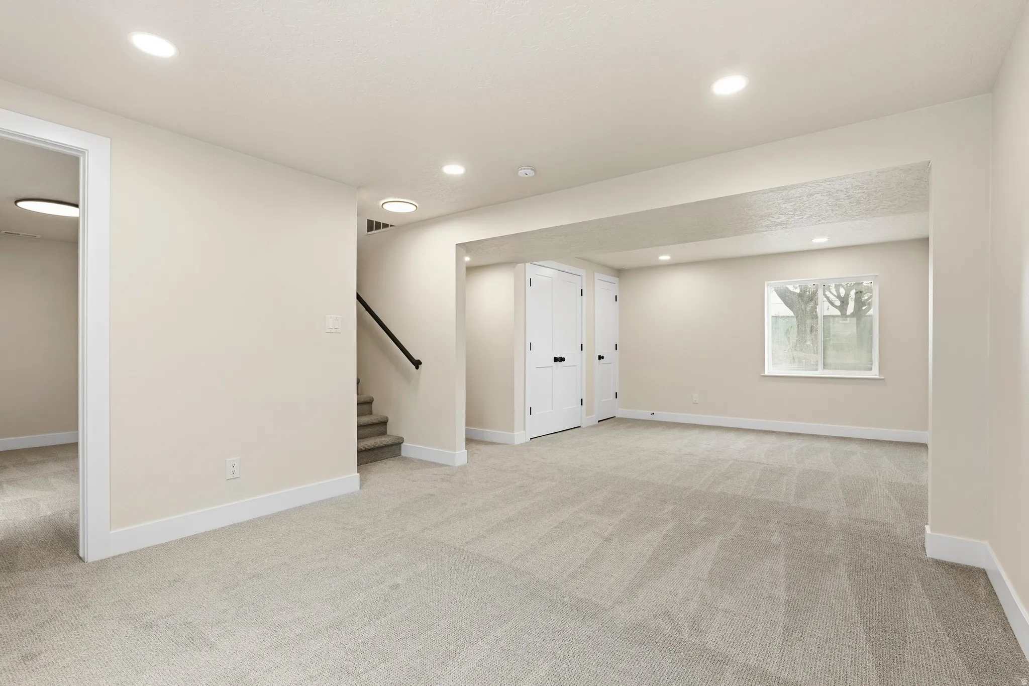 Open-concept room featuring light-toned carpeting, recessed ceiling lighting, a window with exterior tree visibility, and white paneled doors with black hardware