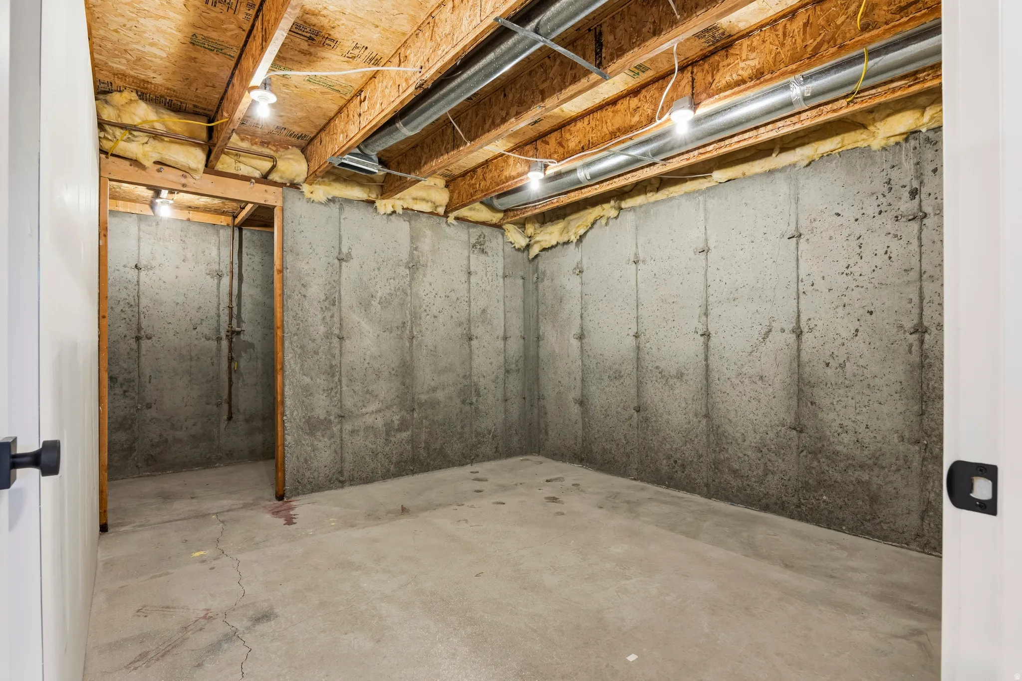 Unfinished basement with exposed wood joists and concrete walls, featuring overhead lighting and visible ductwork
