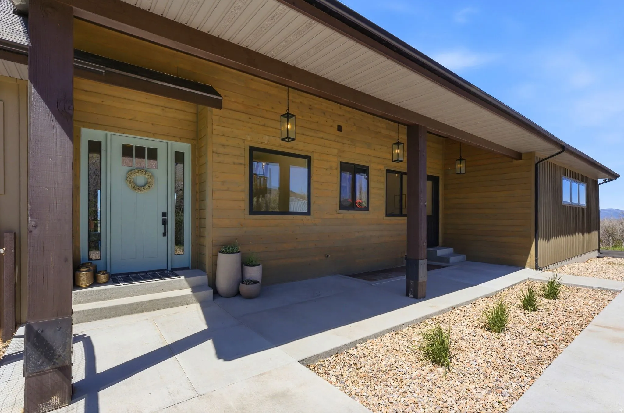 Light blue front door with sidelights and transom window, wood-finish siding, dark framed windows, covered concrete porch, and gravel landscaping