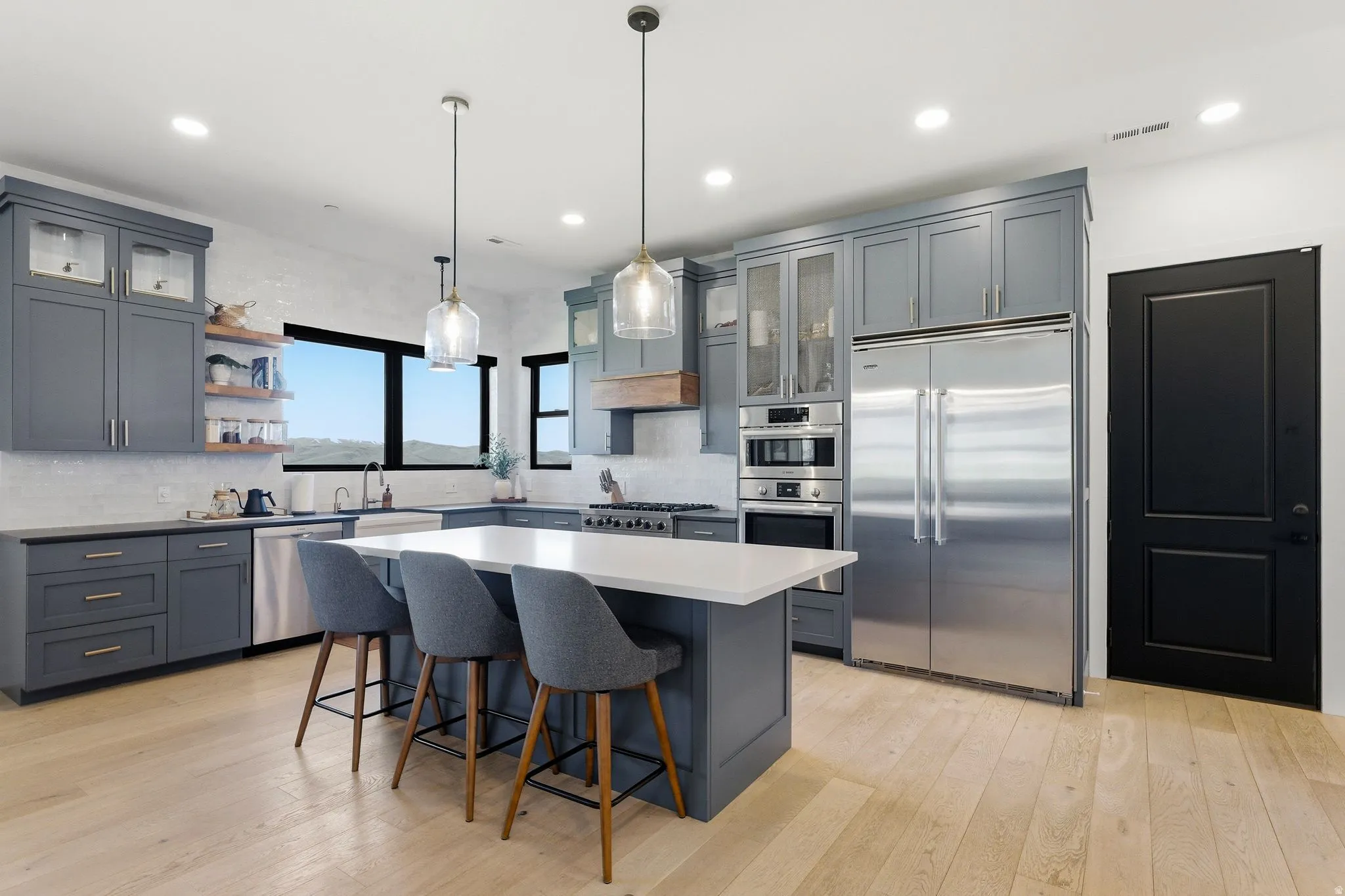 Modern kitchen featuring a large central island with a solid surface countertop, stainless steel appliances, light wood-finish flooring, full height subway tile backsplash, and dark gray cabinetry