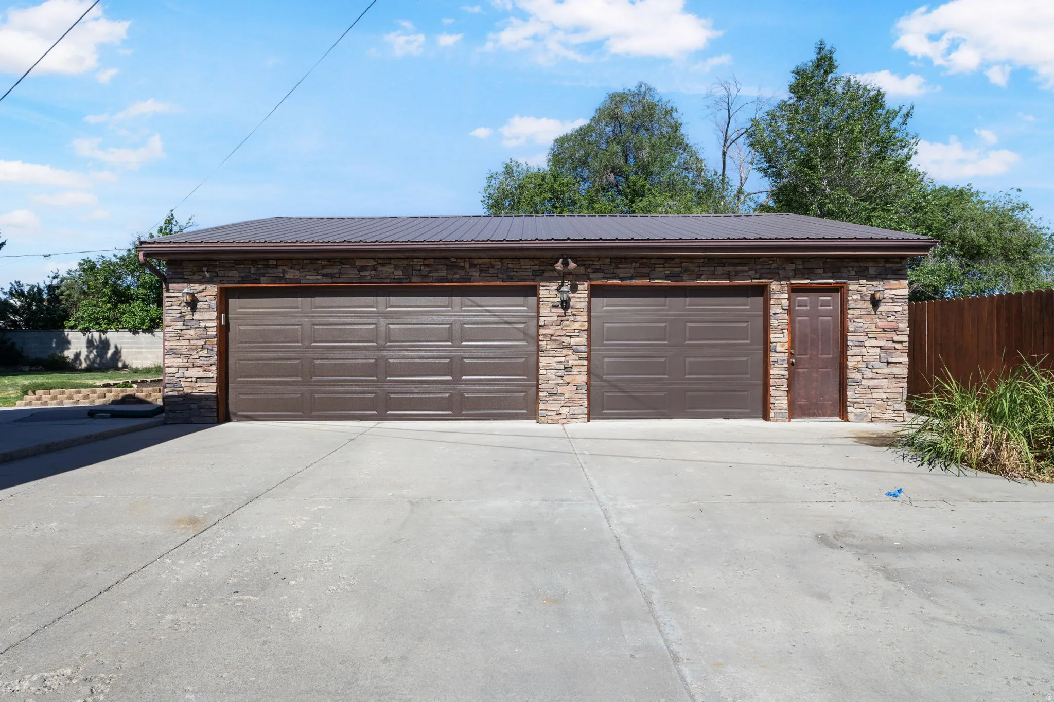 Detached garage featuring three new brown sectional garage doors/openers, a brown metal entry door, and stone veneer siding.