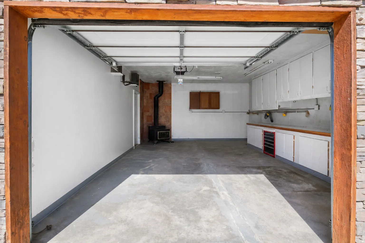 Spacious garage with concrete flooring, stone veneer exterior accents, wood-paneled trim, built-in cabinetry, tool box, workbench, and a wood-burning stove.