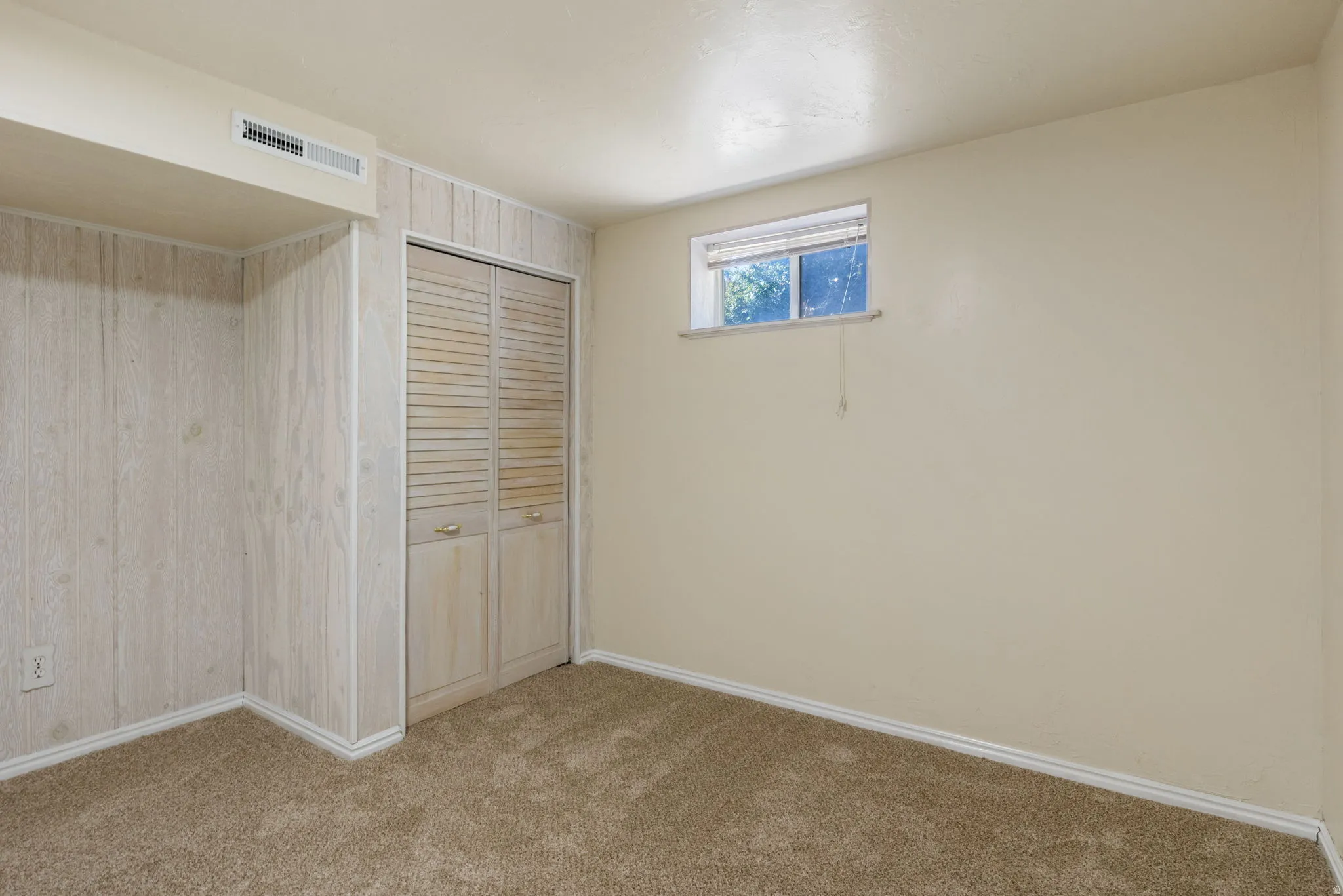 Carpeted room featuring light-toned paneling, a louvered closet, a small window, and an overhead air vent. Basement Bedroom #2-View #2.