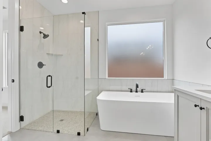 Spacious bathroom featuring a frameless glass shower enclosure, freestanding soaking tub with matte black fixtures, and a vanity with white cabinetry and a light-toned countertop