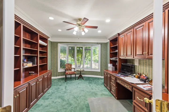 Office space featuring built in desk, wallpapered walls, ornamental molding, dark colored carpet, and a ceiling fan
