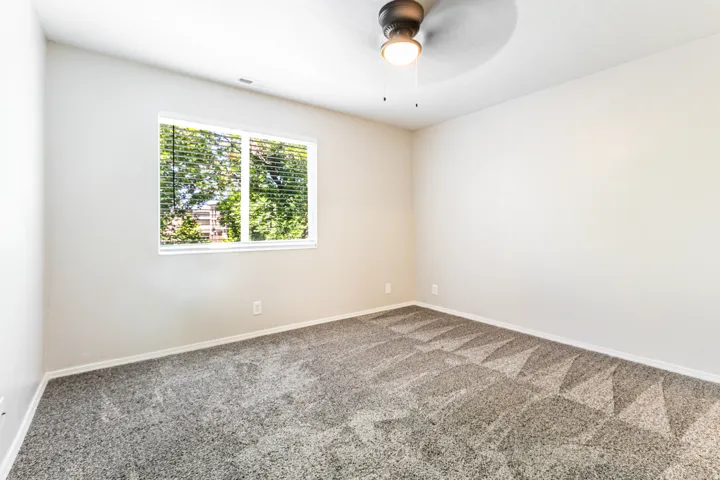 Neutral painted walls defining an expansive room featuring patterned carpeting, a large window with horizontal blinds, and a ceiling fan with integrated lighting