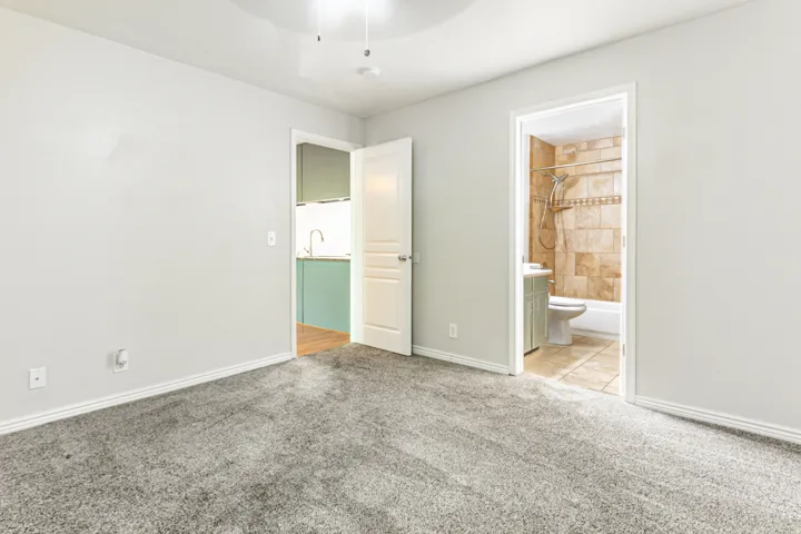 Carpeted room featuring a ceiling fan, white baseboards, and doorways leading to a bathroom with tile flooring and a kitchen with wood-finish flooring