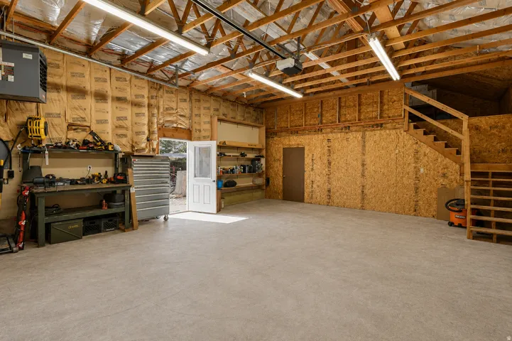 Spacious garage featuring a concrete floor, exposed wood beam ceiling, ample fluorescent lighting, and a built-in wood staircase leading to an upper level