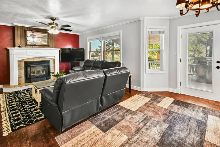 Living area featuring rich wood-finish flooring, a fireplace with a white mantel and stone surround, and an overhead ceiling fan with lighting